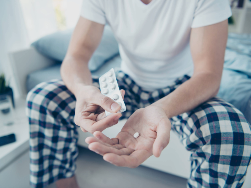 A person in pajama bottoms and a white t-shirt sits on the edge of a bed, holding a blister pack of pills and placing a single tablet into their open hand.