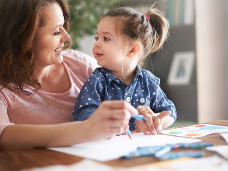 A smiling woman sits at a table with a young child, both facing each other warmly. The child, with hair tied in a small ponytail, holds a crayon while the woman guides them as they draw on paper. Colorful crayons are scattered on the table, and the scene feels cozy and affectionate, suggesting a moment of learning and bonding at home.