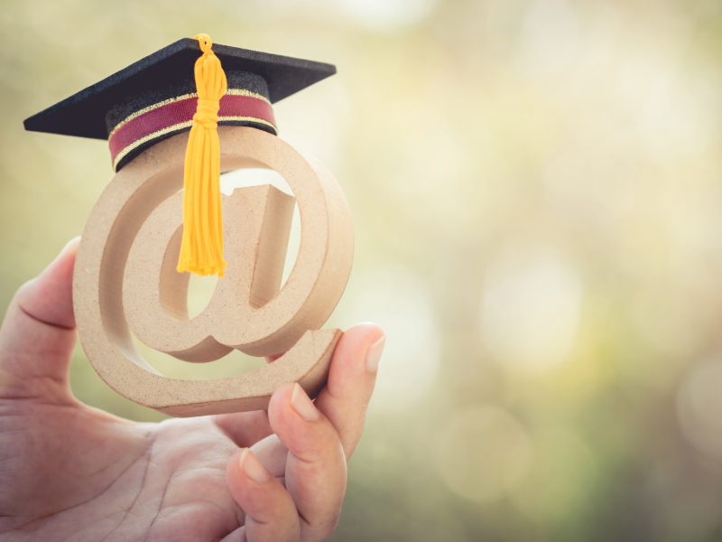 A hand holding a wooden “@” symbol topped with a graduation cap and tassel, symbolizing online education or digital learning.