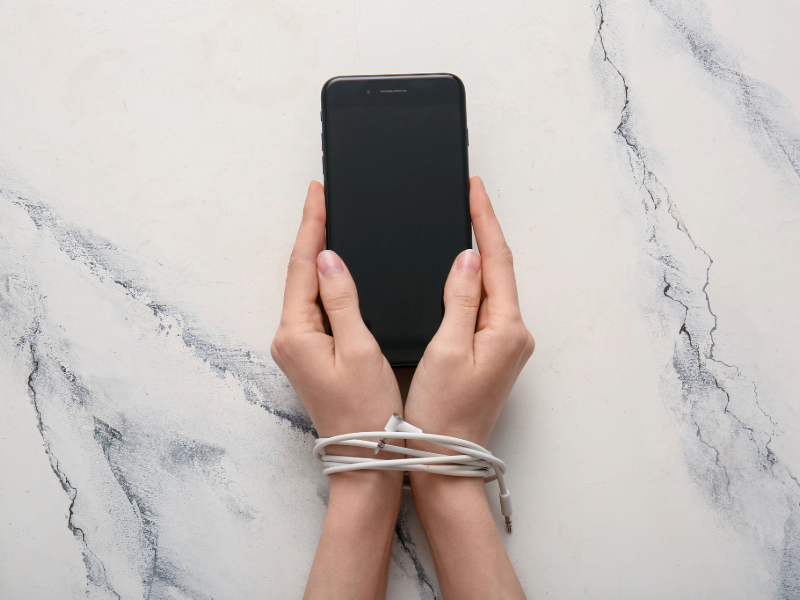 Hands holding a smartphone with a blank screen, wrists loosely wrapped with a white charging cable, symbolizing dependence or restriction related to phone use, set against a light marble background.