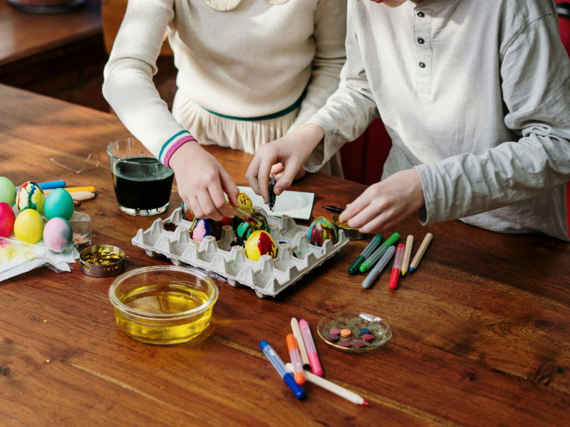 Two children sit at a wooden table decorating colorful Easter eggs. One child carefully paints an egg while the other adds details, with a carton holding several decorated eggs between them. The table is scattered with markers, dye, and small craft supplies, creating a creative and festive atmosphere.