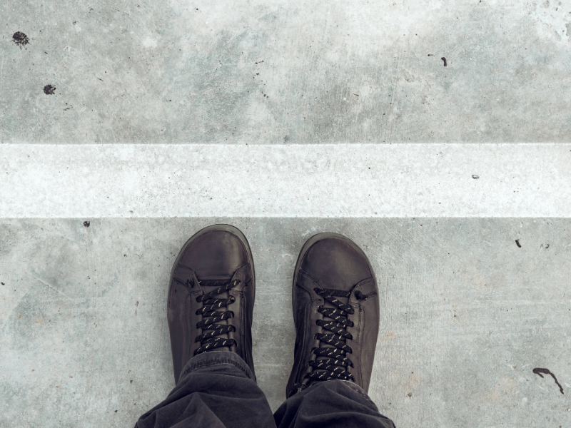 A person wearing black lace-up boots stands on a light grey concrete surface, facing a horizontal white line painted across the ground.