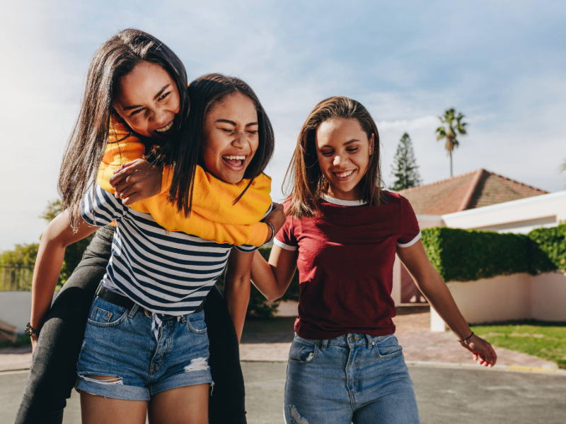 Three teenage girls laughing and having fun outdoors, with one giving another a piggyback ride on a sunny suburban street, capturing friendship, joy, and carefree moments.